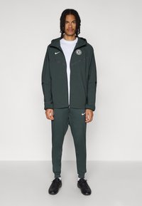 Mørkegrønn Nike joggedress med en glidelåsluejakke med hette, hvit logo, og matchende bukser med elastiske mansjetter og sporty design.