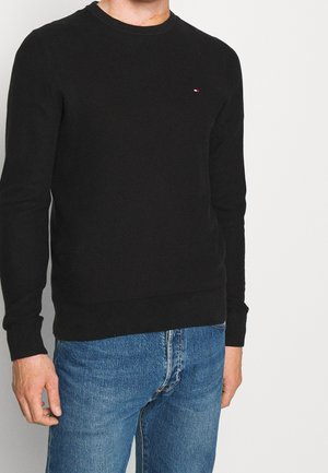 Pull noir en tricot avec un col rond et des poignets côtelés, présentant un petit logo sur la poitrine, porté avec un jean en denim bleu.