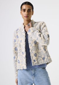 Frau in einer strukturierten blumenartigen Jacke in Creme und Blau über einem blauen Strickpullover und hellblauen Jeans, stehend vor einem schlichten weißen Hintergrund.