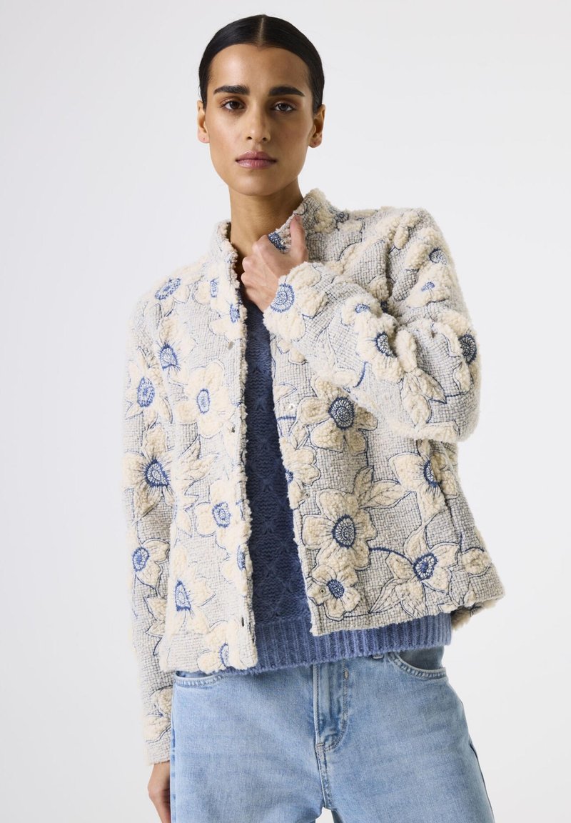 Frau in einer strukturierten blumenartigen Jacke in Creme und Blau über einem blauen Strickpullover und hellblauen Jeans, stehend vor einem schlichten weißen Hintergrund.