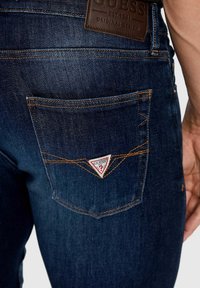 Tummansiniset farkut, joissa on takatasku, jossa on punavalkoinen kolmio-Logo Guess ja kontrastiväriset oranssit ompeleet.