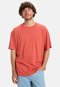 Jonge man met middellang bruin haar, gekleed in een losse koraalkleurige t-shirt en lichtblauwe jeans, staand met één hand in zijn zak.