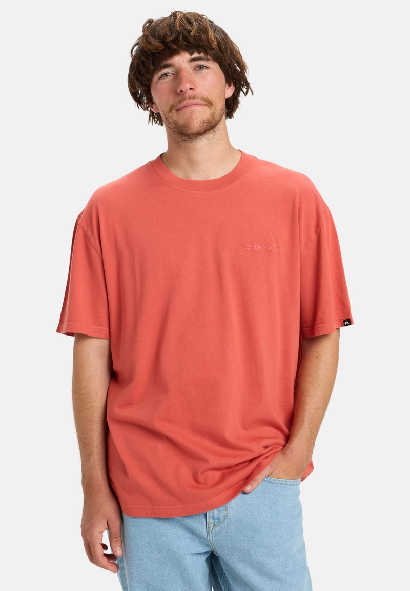 Jonge man met middellang bruin haar, gekleed in een losse koraalkleurige t-shirt en lichtblauwe jeans, staand met één hand in zijn zak.