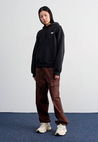 Felpa nera con tasca frontale e logo, abbinata a pantaloni cargo marroni con tasche laterali e sneakers color crema.