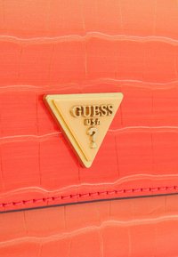 Koralowa torebka ze skóry ekologicznej o fakturowanej powierzchni, ozdobiona złotym trójkątnym logo z napisem "GUESS" oraz detalem z pytajnikiem.