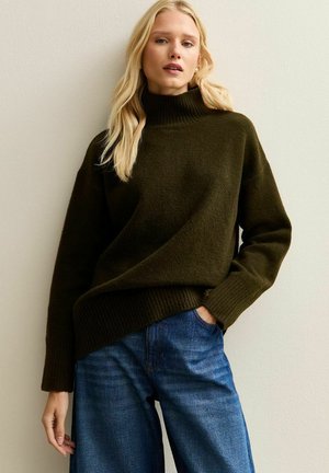 Olivgrüner Rollkragenpullover aus weichem Strickstoff mit gerippten Bündchen und Saum, kombiniert mit lockeren blauen Jeans. Schlichte und entspannte Passform.