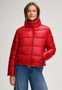 Czerwony puchowy płaszcz z wysokim kołnierzem, z poziomym przeszyciem, zamykanymi kieszeniami i błyszczącą teksturą. Nosi się go na niebieskim dżinsie.