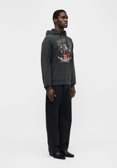 Homme debout portant un sweat à capuche noir avec un motif graphique, un pantalon noir ample et des chaussures noires à bout pointu, sur un fond clair uni.