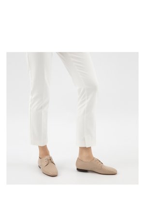 Persoon draagt witte cropped broek en beige geperforeerde veterschoenen met platte zool, staand op een witte ondergrond met één been licht gebogen.