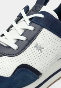 Άσπρο δερμάτινο αθλητικό παπούτσι με τρύπες, με ναυτικό σουέτ διακοσμητικά στοιχεία, άσπρα κορδόνια και ασημένιο λογότυπο. Σχεδιασμός χαμηλής μύτης με υφή λεπτομέρειες.