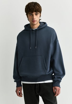 Jonge man met kort bruin haar die een donkerblauwe oversized hoodie en zwarte broek draagt, staand tegen een effen lichte achtergrond.