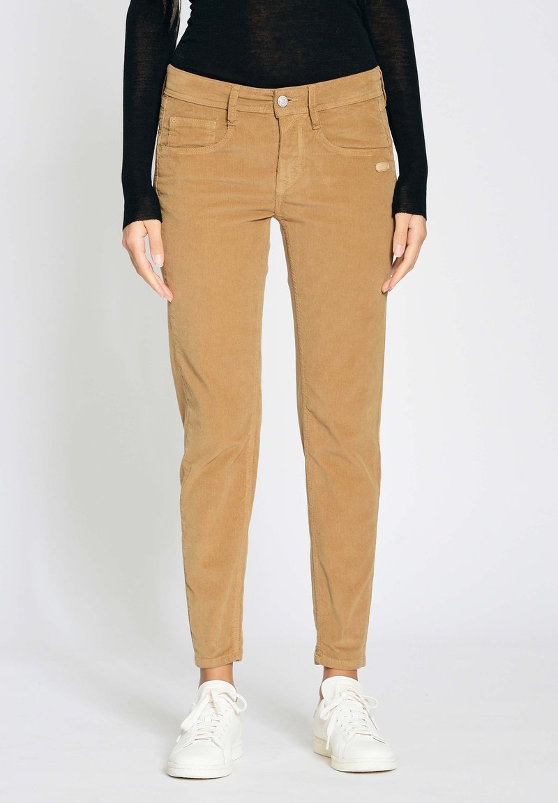 Personne portant un pantalon en velours côtelé beige slim, un haut noir à manches longues et des baskets blanches, debout devant un fond uni.