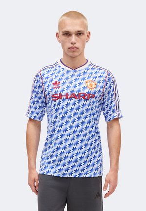 Joven con camiseta de fútbol del Manchester United de color azul y blanco con patrón de zigzag y con el logo rojo "SHARP", de pie frente a un fondo sencillo.