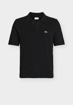 Svart kortermet polo med teksturert stoff, åpen krage og liten grønn Lacoste-krokodillelogo på venstre bryst.