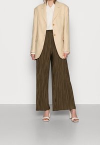 Person wearing a cream blazer, white shirt, olive green wide-leg pleated pants, and white open-toe block heel sandals, standing on a gray floor.