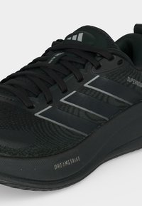 Μαύρο παπούτσι τρεξίματος Adidas με τρεις αντανακλαστικές ρίγες, μαύρα κορδόνια και κείμενο "Supernova" και "Dreamstrike" στην πλευρική σόλα.