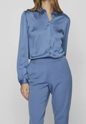 Femme portant un chemisier en satin bleu à manches longues rentré dans un pantalon bleu taille haute et ajusté, sur un fond uni.