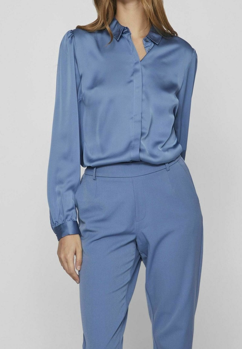 Femme portant un chemisier en satin bleu à manches longues rentré dans un pantalon bleu taille haute et ajusté, sur un fond uni.