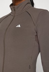 Brun zip-up sportjakke med høj krave, der har diagonale syninger og et lille hvidt logo på brystet. Glat tekstur.