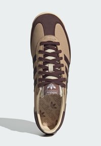 Baskets de sport beige avec des accents bruns, un design perforé et des lacets plats. Comprend un col rembourré et un logo sur la languette et la semelle intérieure.