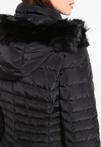 Černá šusťáková bunda s lesklým, prošívaným designem, s lemováním z umělé kožešiny u kapuce a rovný límec. Texturovaná tkanina dodává hloubku.