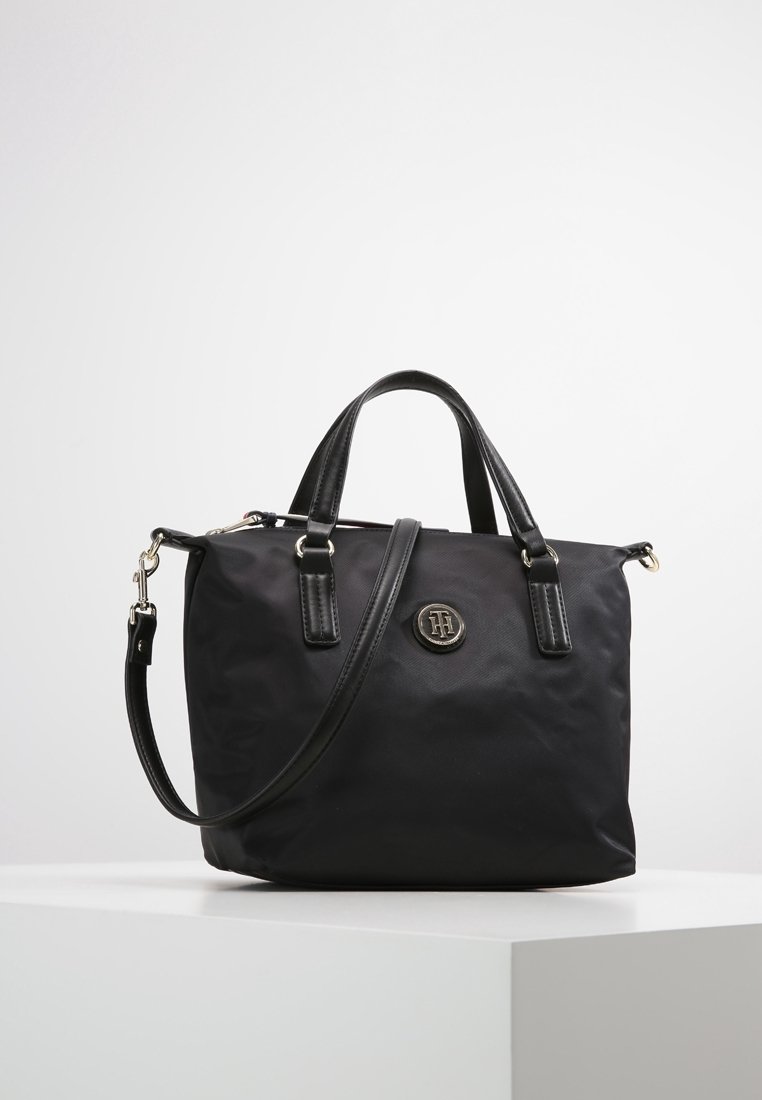Sac à main noir avec doubles poignées et bandoulière réglable, avec un emblème en métal rond du logo au centre devant.