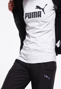 Szary bawełniany t-shirt z czarnym logo "PUMA", noszony pod czarną kurtką na zamek, w połączeniu z czarnymi spodniami z subtelnym detalem logo.