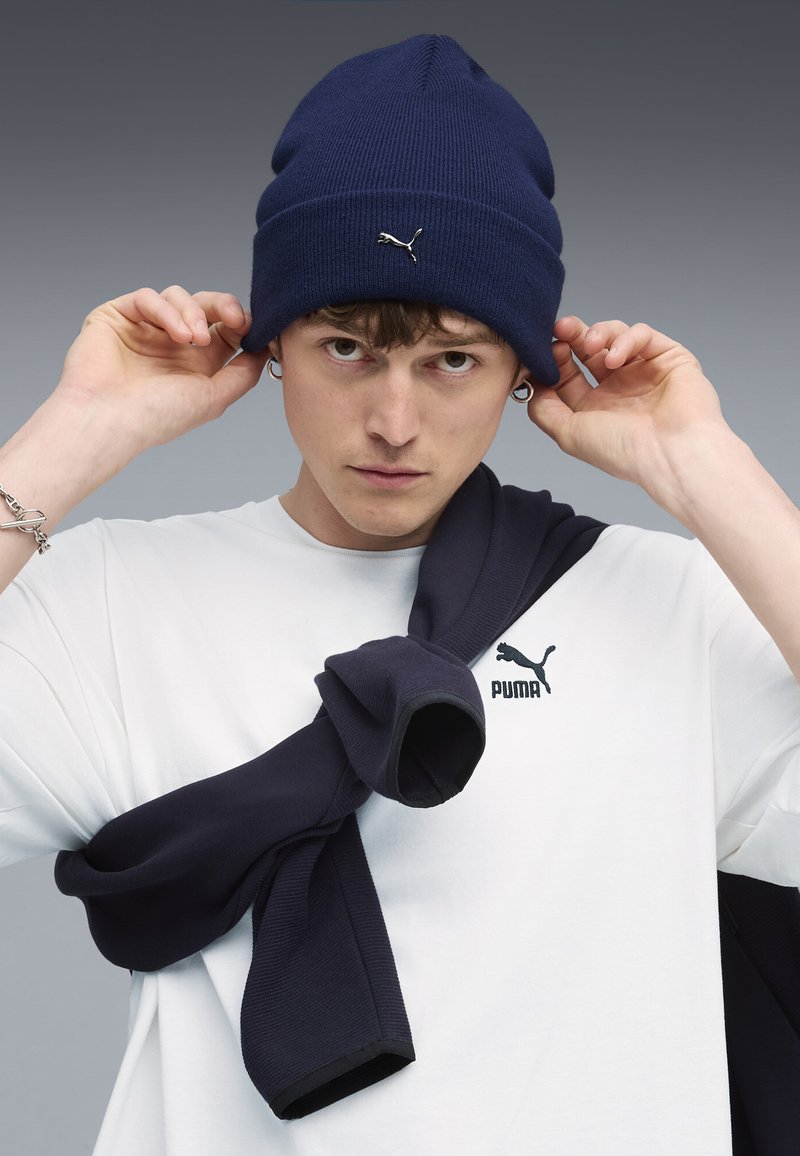 Marineblå beanie med ribbet tekstur, med lille sølvlogo. Modellen har en hvid skjorte på med et sort logo og en marineblå sweater draperet over skuldrene.
