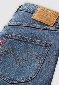 Pantaloni in denim blu con tasca, cuciture arancioni, etichetta rossa Levi's e etichetta in pelle marrone Levi Strauss & Co. sopra il girovita.