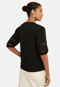 Donna con capelli scuri raccolti in un bun, che indossa una blusa nera con maniche haute in pizzo e pantaloni bianchi a vita alta, rivolta verso destra.