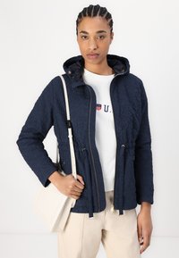 Ναυτικό κουβερτίνα μπουφάν με κουκούλα, διαθέτει ελαστικά κορδόνια, χρυσά εξαρτήματα και φερμουάρ. Φοράει επάνω από ένα λευκό φούτερ.