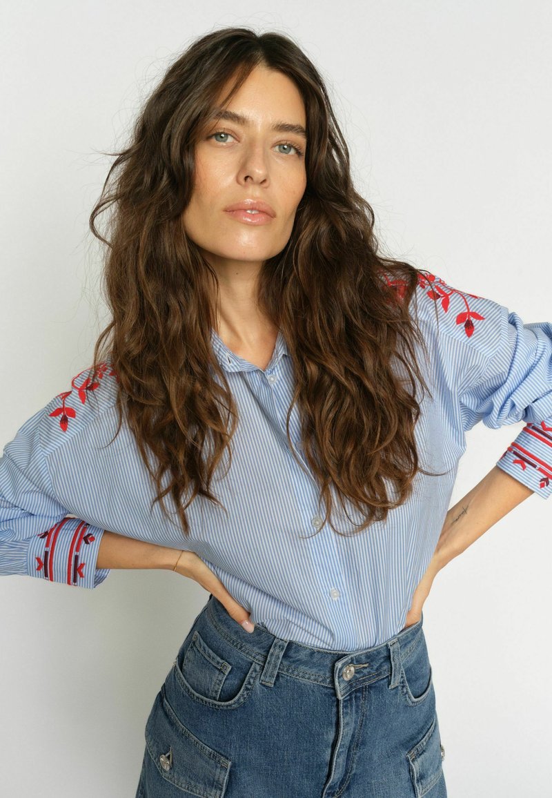 Femme aux longs cheveux bruns ondulés portant une chemise rayée bleue avec une broderie rouge et un jean en denim bleu taille haute, posant les mains sur les hanches.