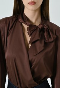 Blusa in satin marrone con fiocco al collo, scollatura a V profonda e maniche drappeggiate, abbinata a una cintura scura.