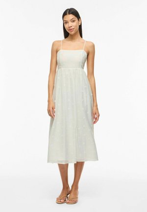 Kvinne står iført en hvit midikjole med tynne stropper og teksturert stoff, kombinert med beige sandaler med hæl på hvit bakgrunn.