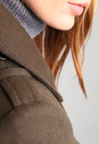 Abrigo de lana verde oliva con un cuello estructurado, costuras visibles y acentos en los hombros. Textura suave, mostrado de lado con cabello.