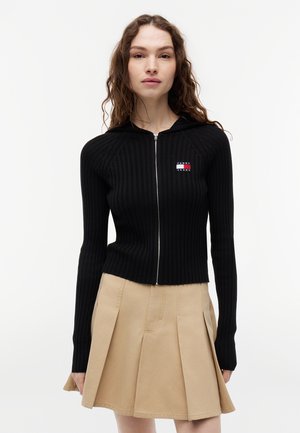 Donna che indossa una felpa con cappuccio nera a coste con zip e piccolo logo con bandiera e gonna beige plissettata, in piedi su sfondo bianco uniforme.