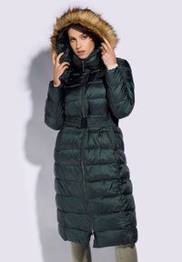 Long puffer coat in dark green, featuring a fur-trimmed hood, cinched waist with a belt, and horizontal stitching across the body.