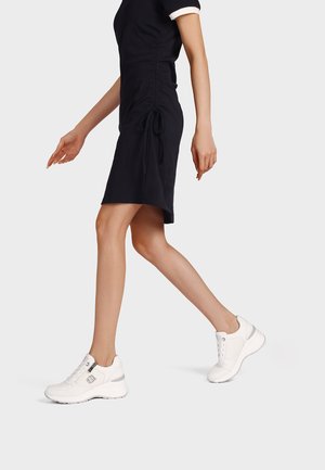 Donna che cammina indossando un vestito nero raccolto ai lati abbinato a sneakers sportive bianche su uno sfondo chiaro e uniforme.