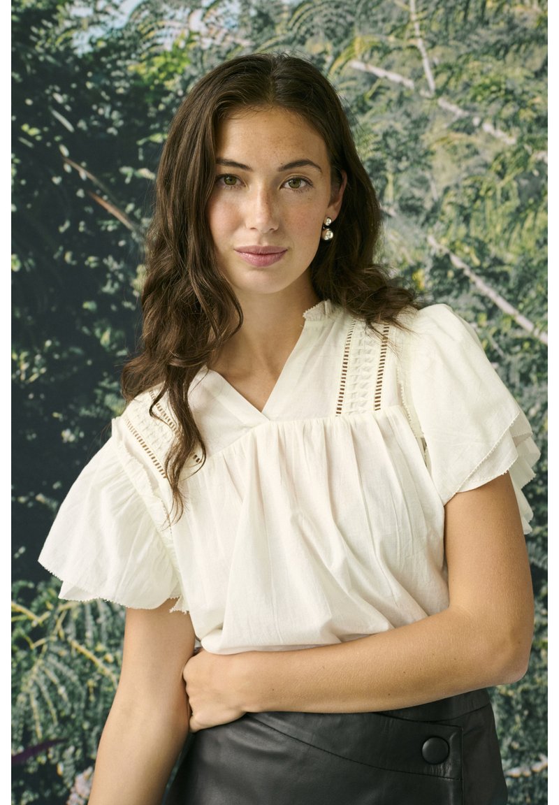 Jeune femme aux longs cheveux foncés portant un chemisier blanc et une jupe noire, debout devant un fond vert feuillu, regardant l'appareil photo.