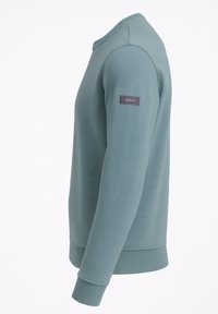 Fleecetröja i mjuk teal med rund hals, långa ärmar, ribbade muddar och en liten varumärkeslapp på vänster ärm.