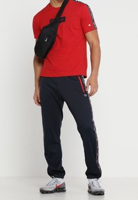 Camiseta roja de manga corta con logo, combinada con pantalones de chándal azul marino que tienen franjas laterales y bolsillos con cremallera. Bolsa cruzada negra. Zapatillas grises.