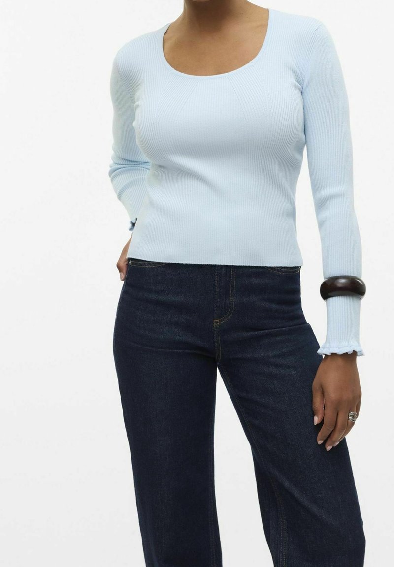 Femme portant un haut à manches longues côtelé bleu clair, un jean bleu foncé, un bracelet noir au poignet gauche et une bague argentée à la main droite.