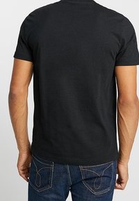 Camiseta de algodón negra con cuello redondo, mangas cortas y una textura suave, combinada con jeans de mezclilla azul oscuro que presentan acentos de costura en contraste.