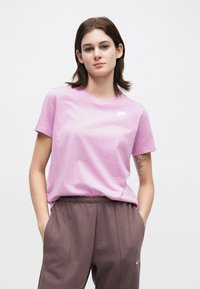 Νεαρή γυναίκα με κοντά καστανά μαλλιά, φορώντας ένα ροζ μπλουζάκι Nike και καφέ φορμάκια Nike, στέκεται με τα χέρια στις τσέπες.