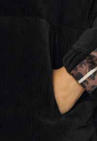Main dans une poche en velours côtelé noir avec un poignet de manche noir portant un tissu imprimé léopard marron et noir en dessous.
