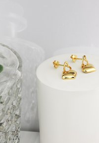 Goldfarbene, herzförmige Ohrstecker mit glänzender Oberfläche und strukturierten Details am Steg, präsentiert auf einer weißen zylindrischen Fläche.