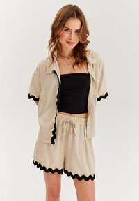 Beige leněná košile a krátké kalhoty s černými vlnitými okraji, krátkými rukávy a černým upnutým tričkem pod nimi. Pas na šňůrku.