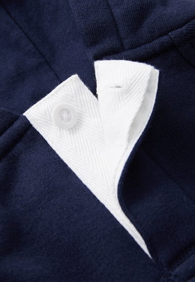 Približan prikaz mornarsko modrega blaga z belim gumbom in teksturirano belo zaponko, ki prikazuje del območja zapenjanja oblačila.