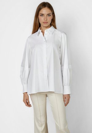Woman with straight light brown hair wears loose white button-up shirt with puffed sleeves and light beige fitted pants, standing against plain background.