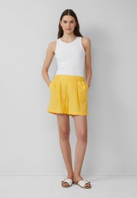 Débardeur blanc, short jaune à plis devant avec poches latérales et sandales blanches avec une accent doré. Texture lisse, coupe décontractée.
